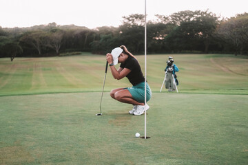 A young woman a beginner golfer, is squatting on the golf course and holding a club in her hands. She is upset because she missed the hole and lost the competition. Frustration from defeat.