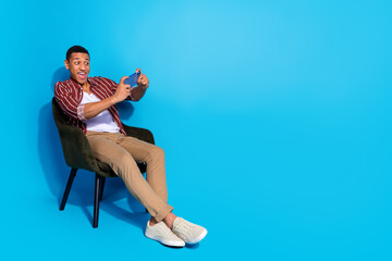Full length photo of funny guy wear stylish t-shirt sit on armchair look at smartphone play video...