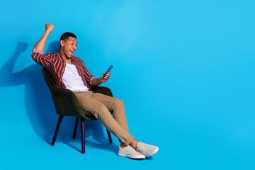 Full length photo of impressed guy wear stylish t-shirt sit on armchair look at smartphone win bet...