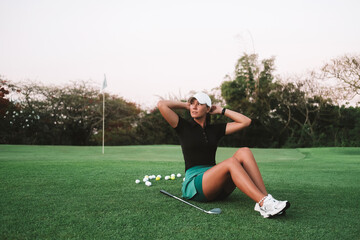 Young attractive woman professional golf coach sitting on a green lawn and posing. Golf lessons at the country club. Portrait of a teacher in a sports uniform. Luxury summer sport.