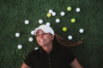 Top view portrait of a young attractive woman in a cap lying on the grass on a green golf course and smiling. Next to her are multicolored balls. Professional player and winner.