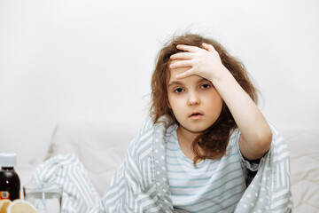 Naklejka premium Sick sad little kid girl sits on bed at home wrapped in blanket and holds her head. Headache. Flu season. Healthcare concept.