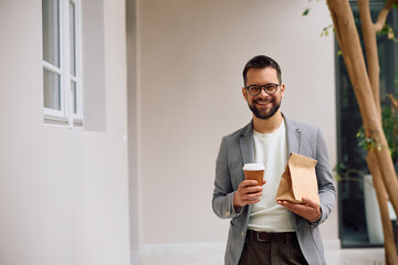 Happy entrepreneur with takeaway coffee and lunch bag in office looking at camera.