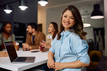 Happy entrepreneur using laptop during coffee break with her colleagues and looking at camera.
