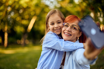 Fototapeta premium A smiling ginger-haired mother takes photos of herself and her daughter while being at the park.