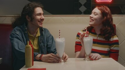 Medium shot of young Caucasian couple laughing while talking during date with milkshakes in retro style cafe at night