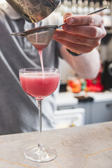 A person pours a pink beverage through a strainer into a stemmed glass on a marble countertop, likely depicting cocktail preparation