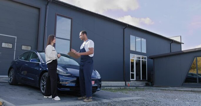 Auto mechanic give customer discussion on her vehicle repairs problems condition outdoor with repair garage station on background. Customer and Automotive Technician Discuss Car Problem