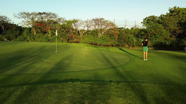 Aerial view of a young woman playing golf on the green lawn at the club. She hits the ball with her club and hits the hole. The woman sits down and jumps with happiness. Hitting the target.