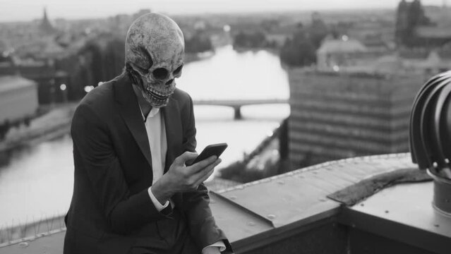 Addicted man sitting on a rooftop in a skeleton mask, using his phone.