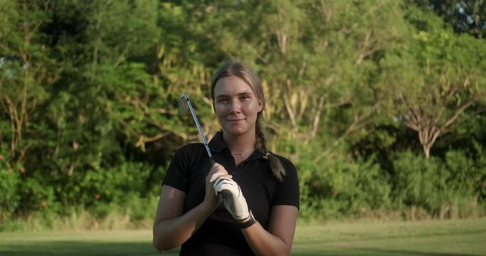 Portrait of a young attractive woman who plays golf professionally on a green lawn. She is holding a golf club in her hands and looking at the camera smiling. Luxurious summer sport.