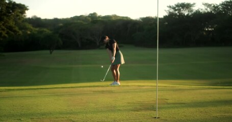 Focus on a golf hole in the background, out of focus a young aspiring female golfer is trying to hit the ball into the hole. She swings and hits on the ball but misses, a failure in the game.