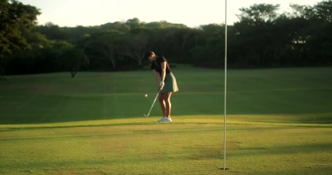 Focus on a golf hole in the background, out of focus a young aspiring female golfer is trying to hit the ball into the hole. She swings and hits on the ball but misses, a failure in the game.