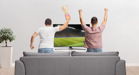 Rear view shot of young men watching football game on tv and jumping from a sofa