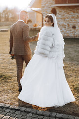 A bride and groom are walking down a path in front of a building. The bride is wearing a fur coat and a veil