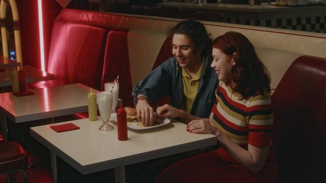 Side shot of young Caucasian couple eating fries and enjoying time during date in 1970s style American diner in evening
