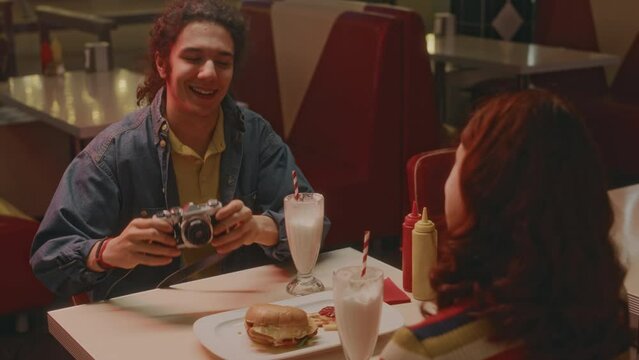 Rear shot of unidentified red haired girl sitting on date with talkative Caucasian guy who taking camera shots in neon lit 1970s style American diner