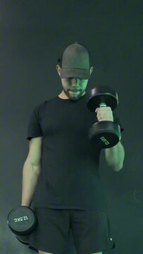 Hombre joven en forma haciendo pesas con gorra frente a una pared de color negro.