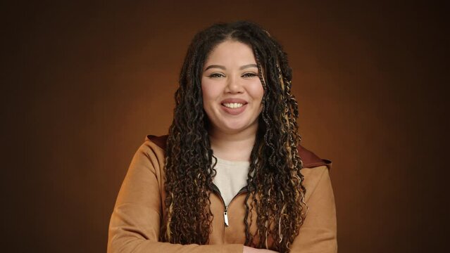 A cheerful young woman with curly hair and a casual jacket smiles warmly against a brown background, exuding confidence and happiness. Perfect for portraying a casual lifestyle theme