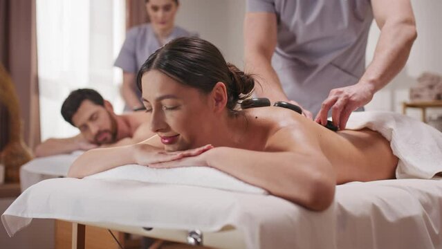 Happy couple relaxing in spa on their honeymoon. Young people receiving professional massage. Skilled massagist stroking back with hot stones. Resting together in wellness center. Resort concept.