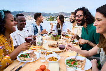 Gathering of happy people around meal table with wine glasses and food celebration terrace barbecue party. Young multiracial friends enjoying laughing together lunch time on summer weekend on rooftop