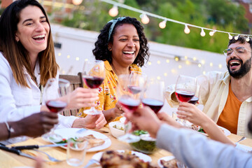 Meeting of joyful young friends toasting with red wineglass celebrating summer party on terrace at home. Happy people enjoying laughing together drinking alcoholic beverages sitting at food table
