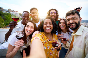 Selfie group of multiracial smiling friends enjoying at rooftop summer party holding glass of red wine. Happy community young people taking picture drinking alcoholic beverages on sunny day together