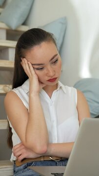 Tired woman listens to boring online conference via modern laptop. Young student sits on wooden stairs at home putting hand on head Vertical Shot.
