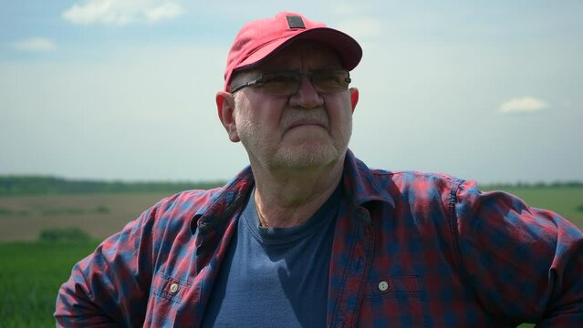 An elderly farmer in a plaid shirt and a red baseball cap stands in a field and surveys the landscape against the blue sky. Slow motion.