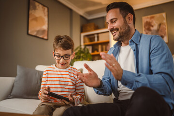 Father and son sit on sofa and use mobile phone at home have fun