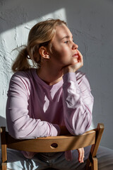 A pensive teenage girl in a pink jacket sits against a wall in the sunlight and looks to the side.