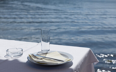  Table set for dinner with glass, plate, cutlery and ashtray on white tablecloth overlooking calm sea. Copy space.food and travel content, especially for seaside restaurants and coastal dining.