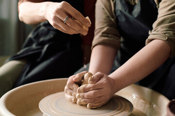 Hand of unrecognizable potter adding water onto clay while student shaping clay on wheel