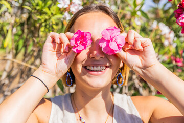 A woman is holding a pink flower in each eye and smiling. Concept of playfulness and joy - Powered by Adobe