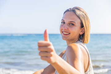 A redhead young woman is smiling and giving a thumbs up while standing on a beach