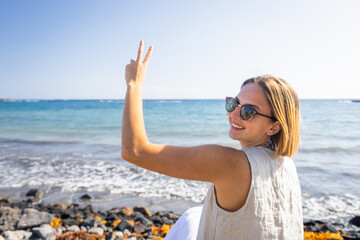 A woman is sitting on a beach with her arm lifted and a peace sign on her hand