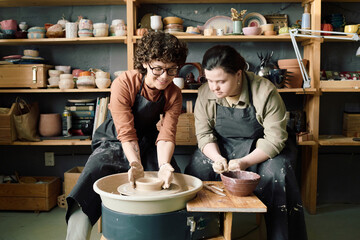 Mature Caucasian artisan demonstrating young female student with Down syndrome how to shape clay on potters wheel during class