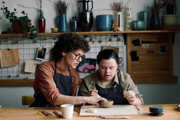 Mature female potter sitting at table teaching young woman with Down syndrome how to make ceramic...