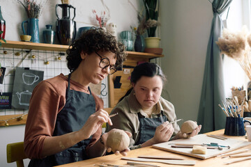 Mature female artisan sitting at table in pottery workshop teaching young woman with Down syndrome...
