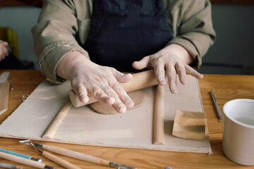 Hands of unrecognizable woman sitting at table in workshop rolling out piece of clay with rolling pin