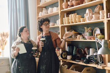 Two diverse female artisans wearing aprons holding cups with coffee drinks looking at earthenware on shelves and discussing work