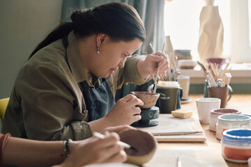 Side view of young woman with Down syndrome sitting at table in pottery workshop painting ceramic...