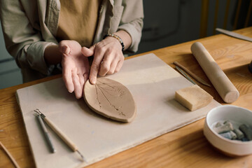 High angle view of hands of unrecognizable artisan sitting at table shaping clay leaf plate