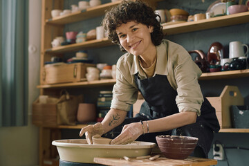Portrait of cheerful female artisan wearing apron smiling at camera while shaping bowl on potters wheel - Powered by Adobe