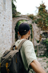 Follow me concept. A young man with a retro-style green backpack travels in old town Bar in Montenegro. Rear view. Stari Bar - ruined medieval fortress in mountains, Unesco World Heritage Site.