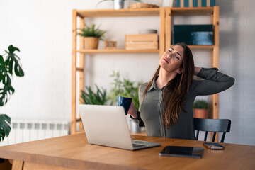 Young self-employed woman sitting in her home office and stretching in chair after working on...