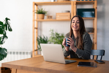 Happy charming female entrepreneur on break holding coffee cup while working over laptop from home.