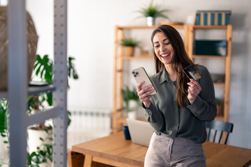 Happy young woman holding credit card using cell phone mobile app paying online making purchase in...