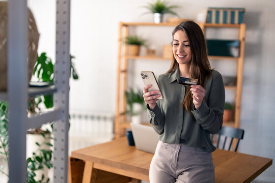Young businesswoman consumer holding credit card leaning on desk at home doing online banking transaction over smart phone. E-commerce virtual shopping, secure mobile banking concept.