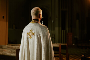 Rear view of unrecognizable elderly Catholic priest wearing vestment standing in empty church, copy space